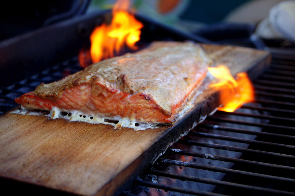 A wild BC salmon filet dressed with Denzel's White Wine Dill Mustard on a cedar plank sitting on a bbq grill with flames rising from both sides. Looking absolutely succulent. 